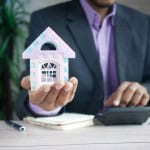 A man in a suit holding a toy house and typing on a calculator to determine if capital gains taxes need to be paid on a house sale